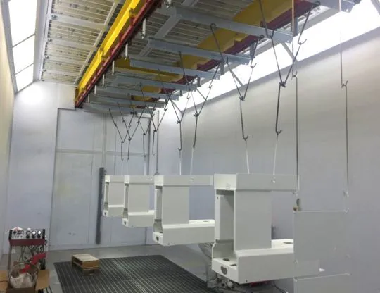 Vista interna della cabina di verniciatura a polveri pressurizzata con pezzi appesi alle barre trasversali del convogliatore aereo